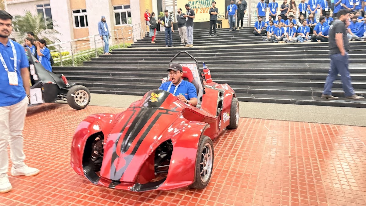 Driver posing with a red REEV vehicle on the GITAM University steps in front of a crowd of student volunteers